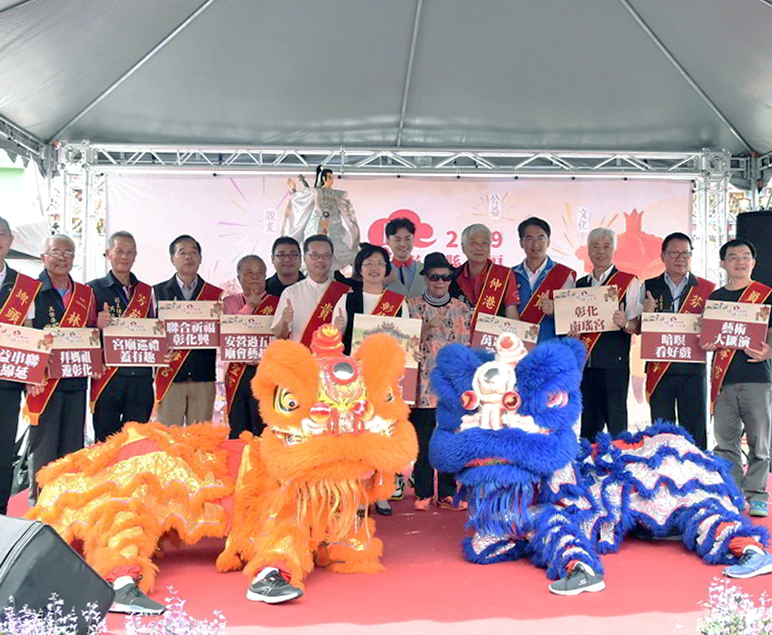 무대 위에 사람들이 단체로 서 있고, 앞쪽에는 주황색과 파란색 사자탈 인형이 놓여 있는 행사 장면