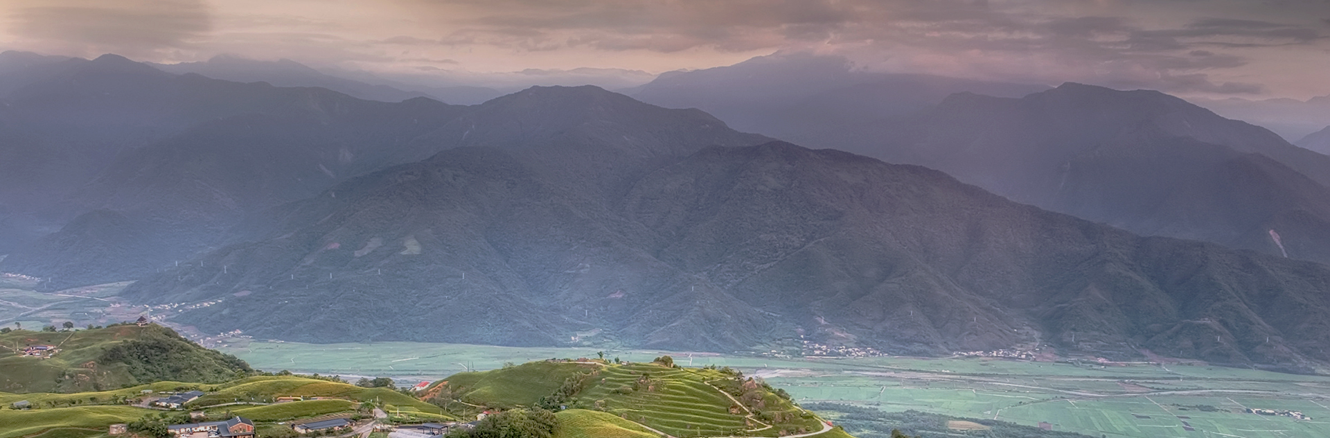 겹겹이 이어진 산맥과 초록빛 들판이 어우러진 풍경, 흐린 하늘 아래 구름이 산 능선을 덮고 있는 모습