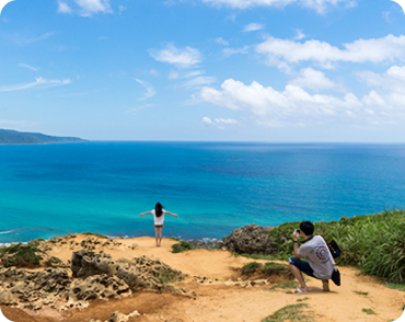 푸른 바다와 하늘이 펼쳐진 해안 절벽 위, 한 사람이 바다를 향해 두 팔을 벌리고 서 있고 다른 사람이 그 모습을 카메라로 촬영하고 있다。